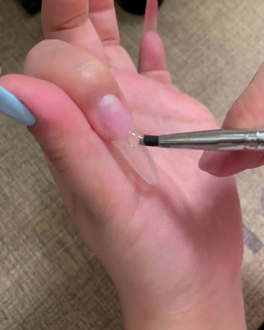 Un pincel aplicando una capa de esmalte en gel rosa nude sobre una uña preparada.