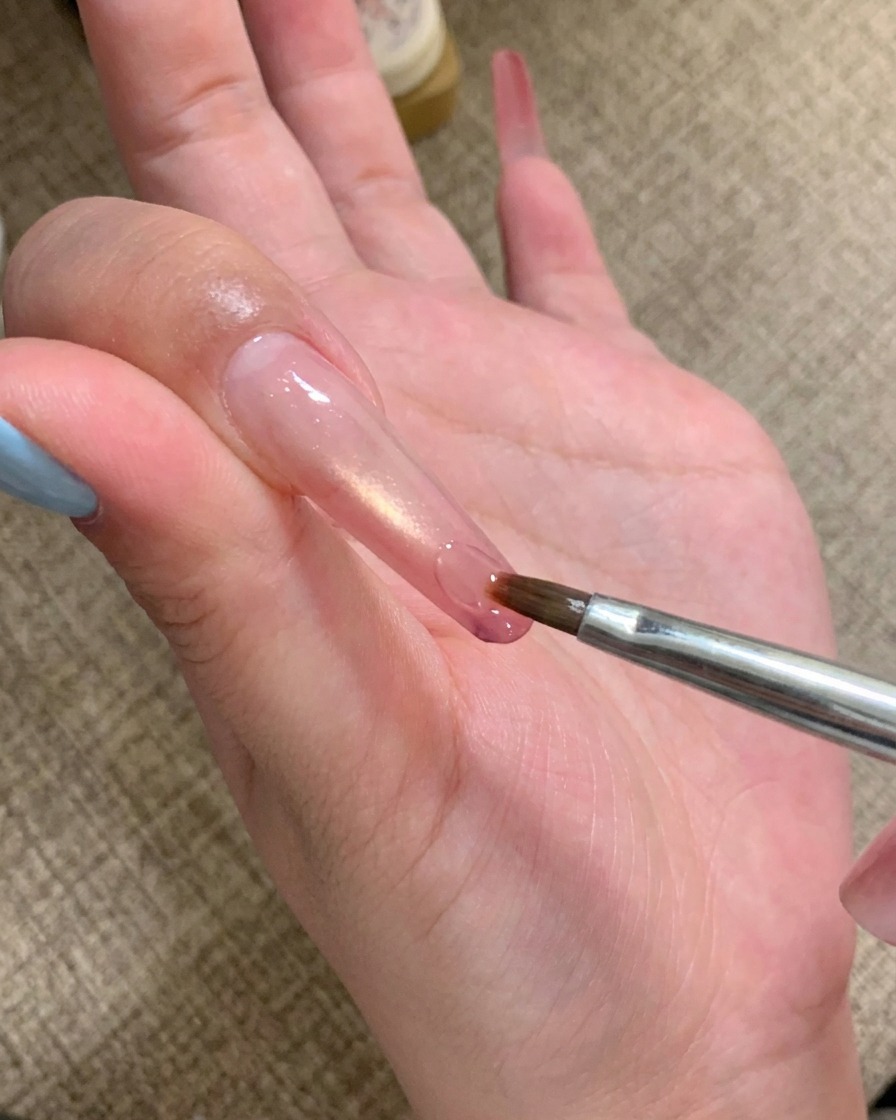 Un pincel de nail art aplicando una gota de gel adhesivo transparente en la punta de una uña larga.