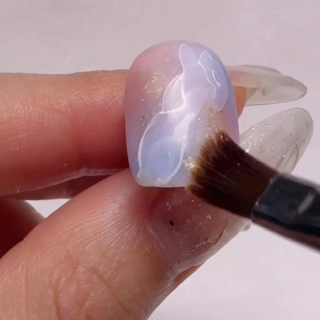 Un primer plano de un pequeño pincel de uñas usando alcohol para difuminar y suavizar suavemente un borde del nail art blanco sobre una base degradada rosa y azul.