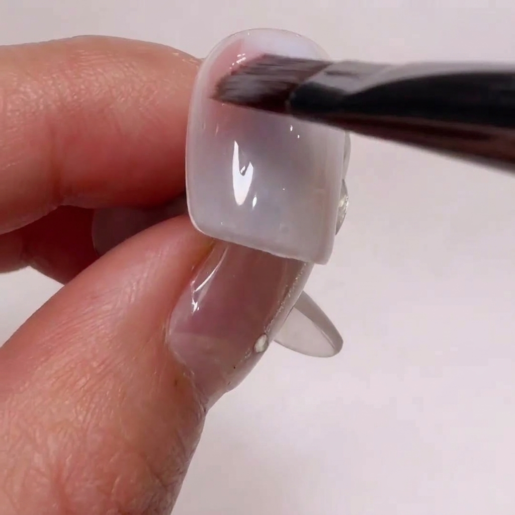Un pincel de nail art aplicando un gel rosa nude suave en las esquinas laterales de un tip de uña.