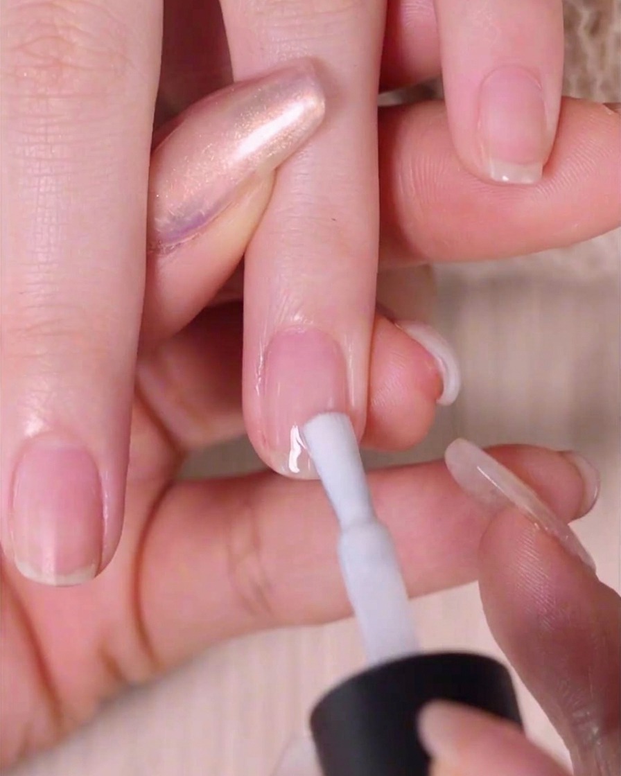 Primer plano de una mano usando un pincel para aplicar una capa fina de esmalte blanco transparente sobre una uña natural.