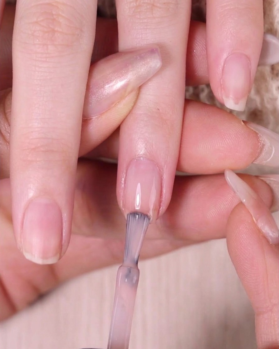 Un pincel aplicando esmalte rosa nude transparente estrictamente en el tercio superior de la uña.