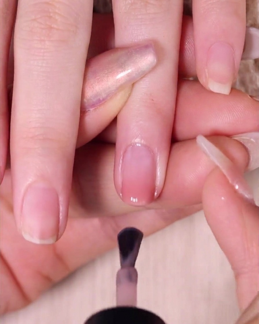 Un pincel aplicando una capa suave de esmalte rosa nude transparente sobre una uña ombré, dejando un pequeño espacio cerca de la cutícula.