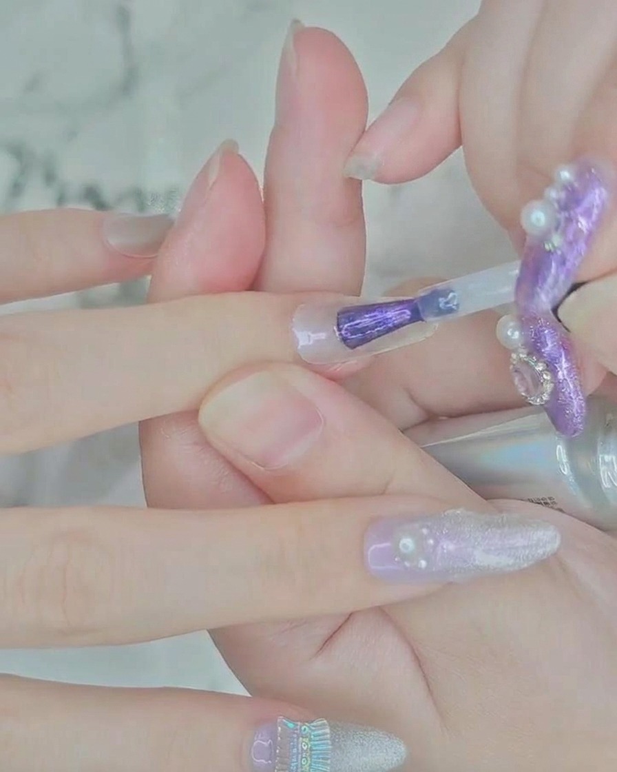 Primer plano de un pincel aplicando esmalte en gel morado translúcido en el centro de una extensión de uña transparente.