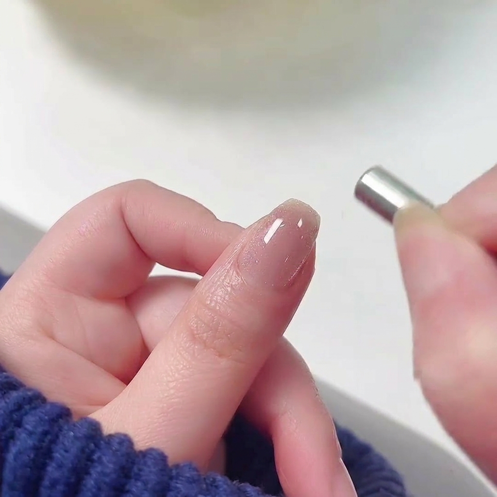 Un pequeño imán cilíndrico siendo sostenido cerca de la cutícula de una uña cubierta con esmalte gel cat-eye rosa brillante y húmedo.