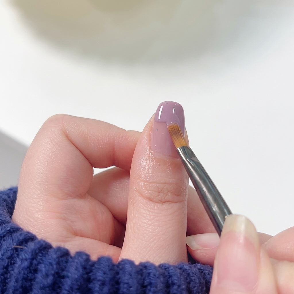 Un pincel plano para nail art barriendo suavemente el borde inferior del esmalte morado hacia la base rosa para crear un degradado.