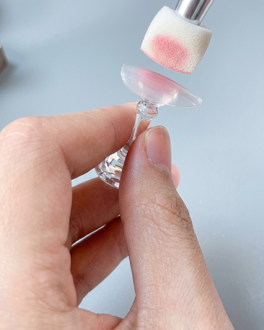 Primer plano de una esponja aplicando un suave color rosa rubor en un tip de uña sobre un soporte.