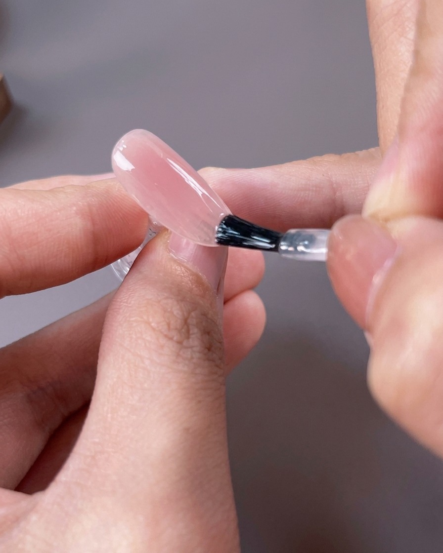 Un pincel aplicando un esmalte de gel translúcido en tono nude rosado sobre toda la superficie de la uña.