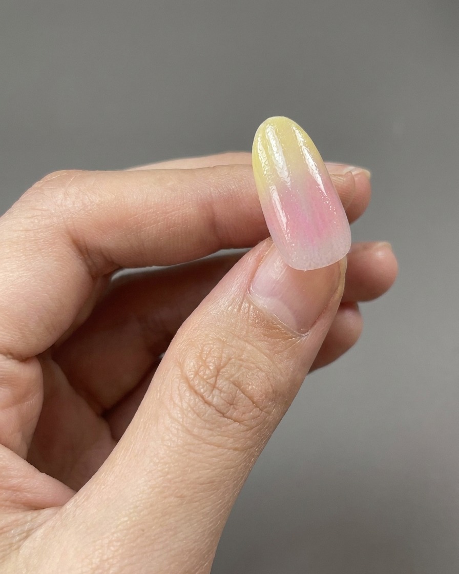 Un pincel aplicando una capa base transparente y brillante sobre un diseño de uñas degradado en tonos amarillo y rosa.