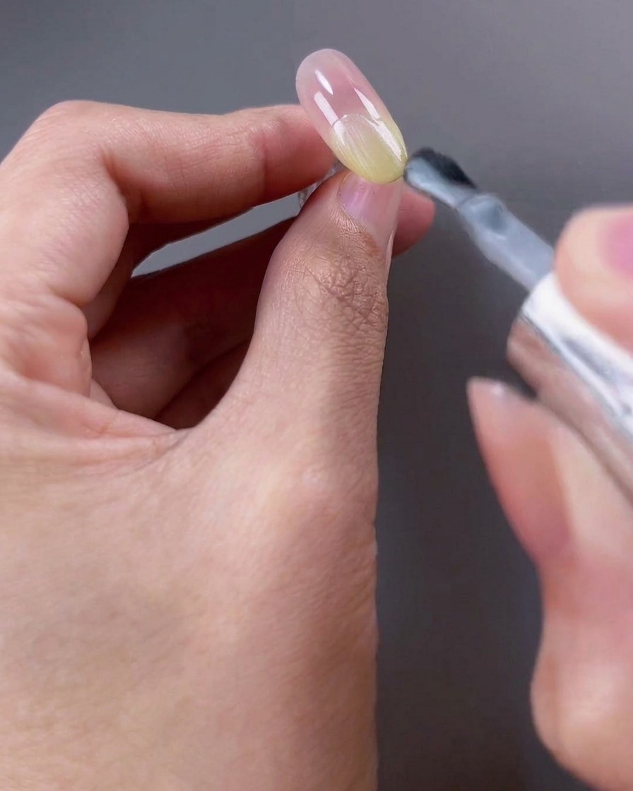 Un pincel aplicando esmalte de gel blanco brillante translúcido en la mitad superior de la superficie de la uña.