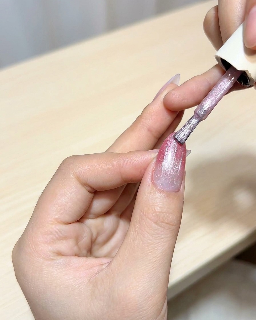 Un pincel aplicando una capa de esmalte en gel ojo de gato sobre una base ombré magenta.