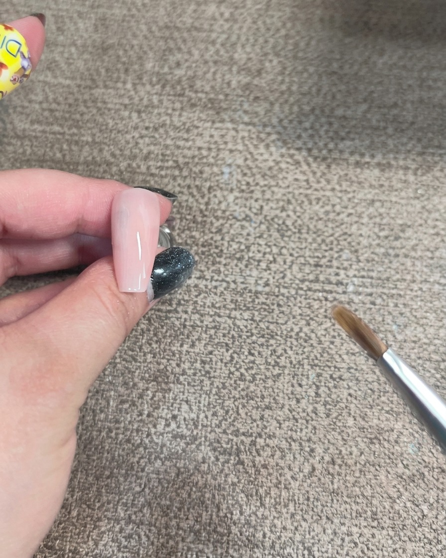 Primer plano de un pincel aplicando gel iridiscente en curva sobre una base de uña rosa claro.