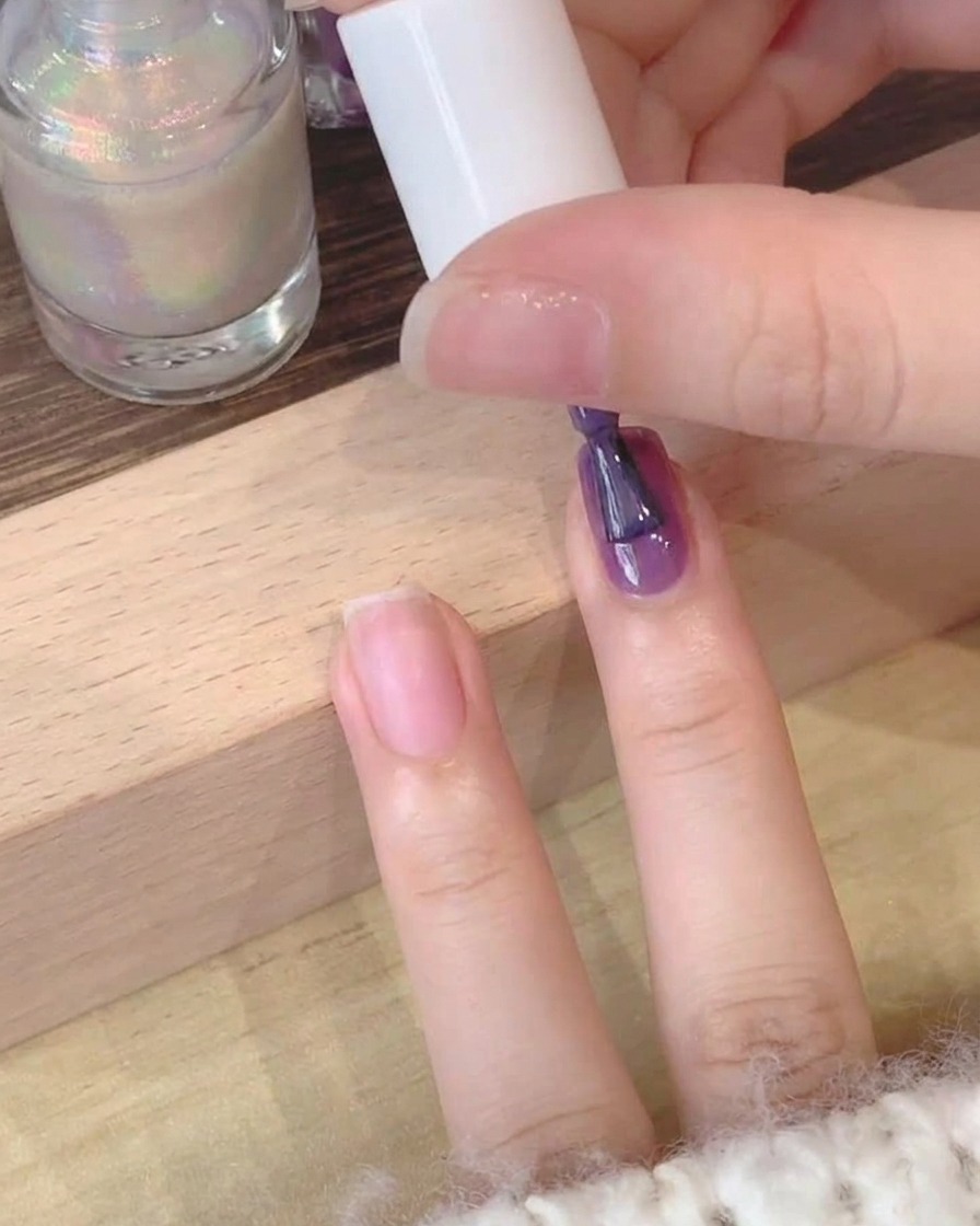Vista en primer plano de un pincel aplicando esmalte de gel púrpura oscuro sobre una uña natural.