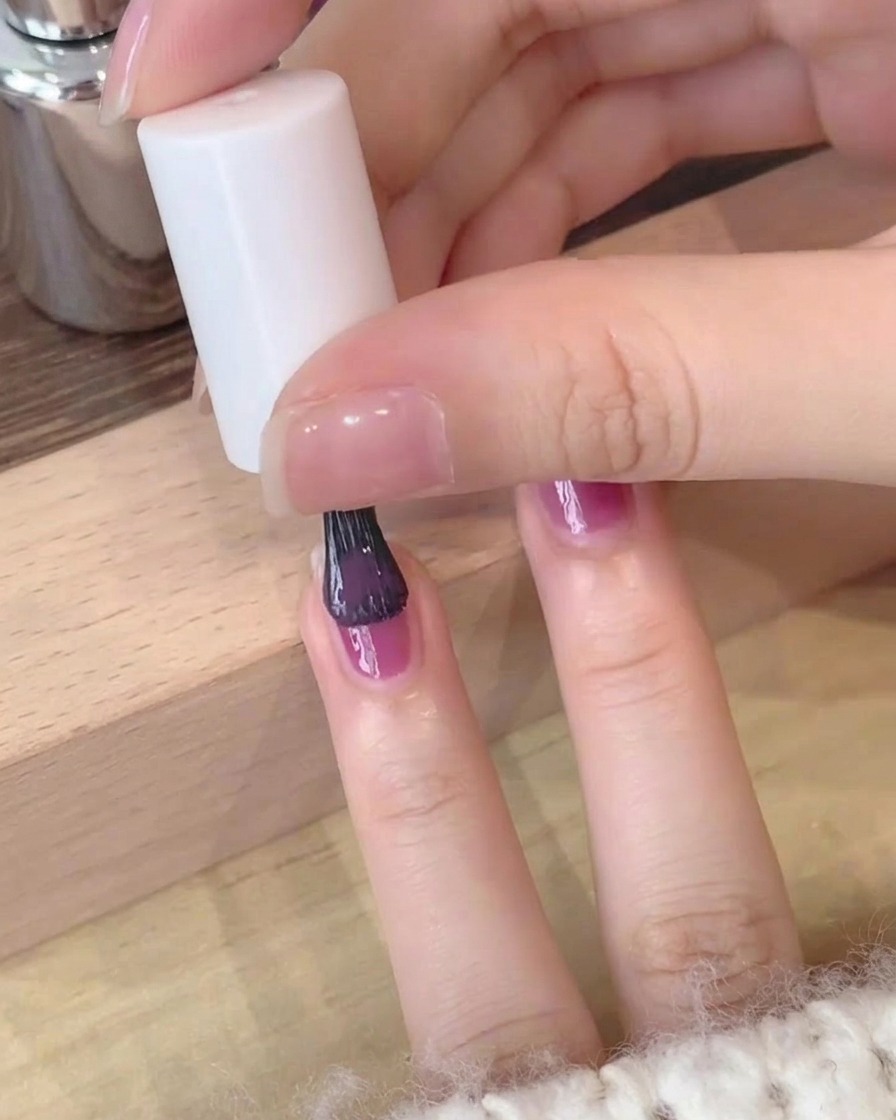 Primer plano de una segunda capa de esmalte de gel púrpura profundo siendo aplicado cuidadosamente sobre una uña.