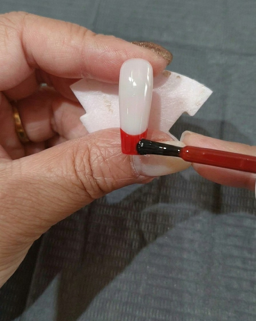 Una capa fina de esmalte de gel rojo siendo aplicada en la parte inferior de una uña con base negra.
