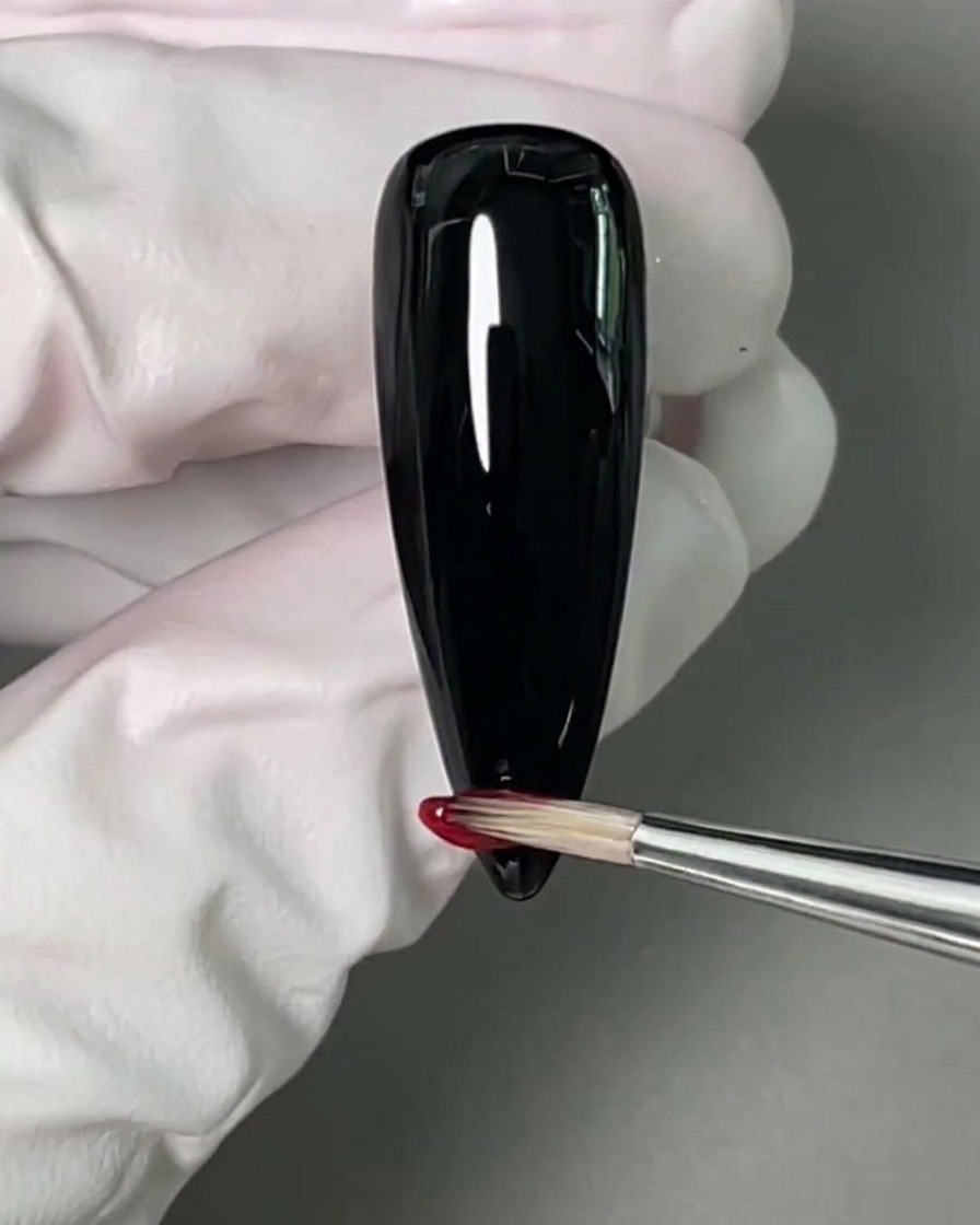 Un pincel fino aplicando esmalte en gel rojo en la punta de una uña pintada de negro.