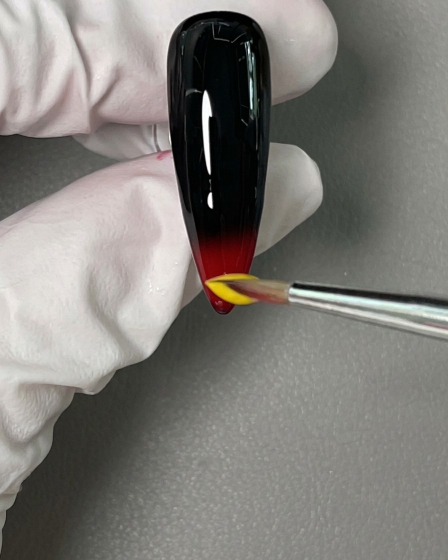 Un pincel de detalle colocando una gota de esmalte amarillo sobre el gel rojo húmedo en la punta.