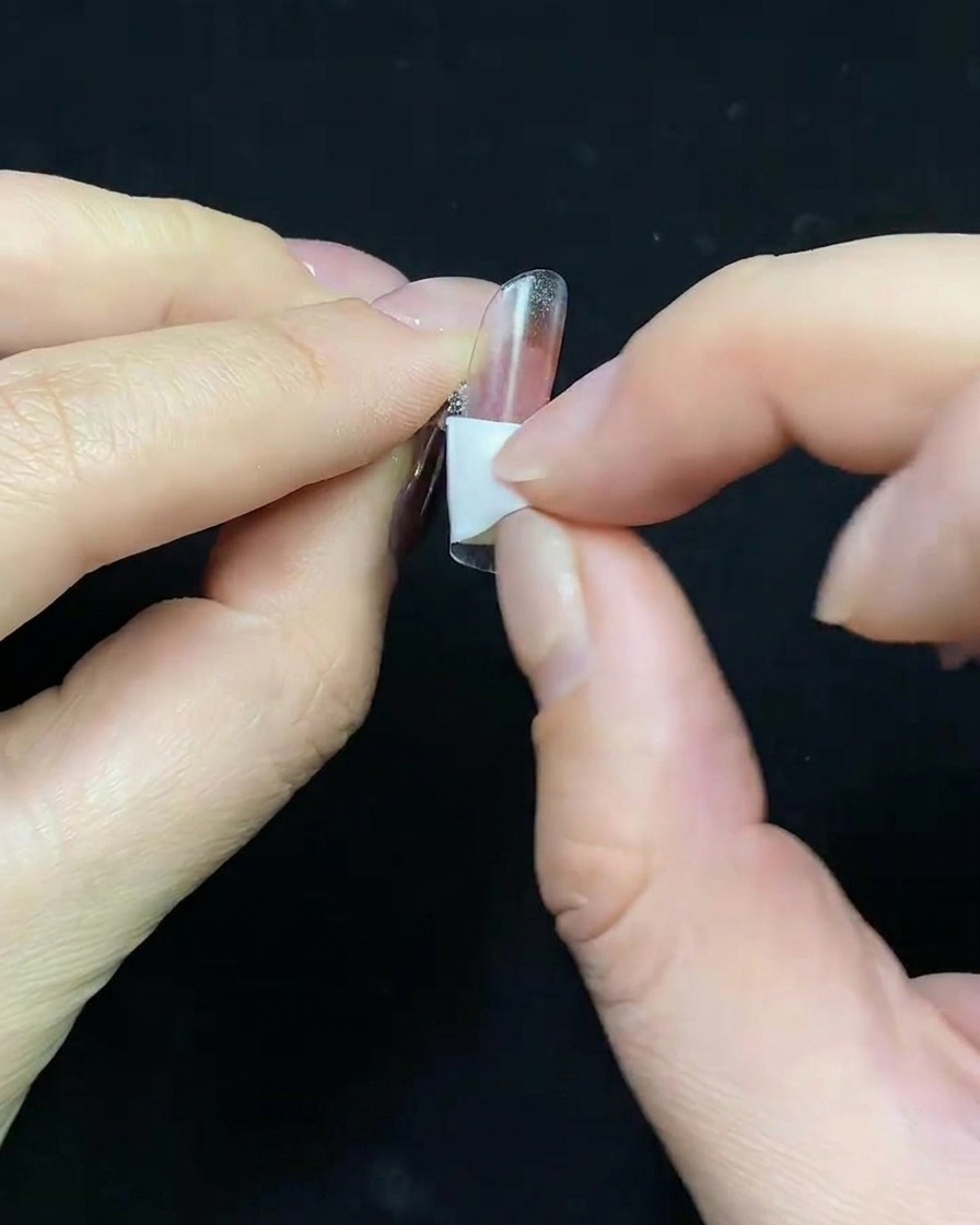 Primer plano de un pincel aplicando base gel color nude sobre un tip de uña transparente.