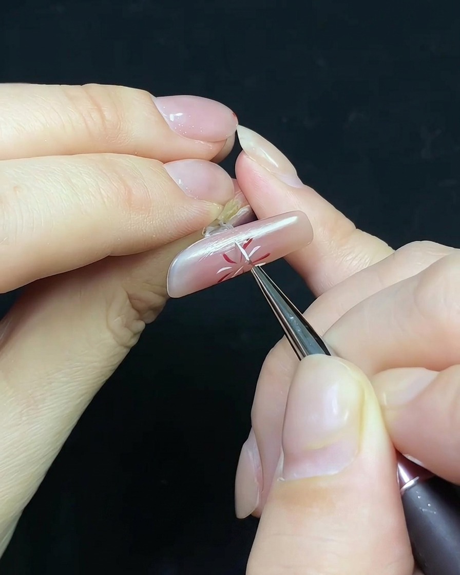 Un pincel fino aplicando top coat sobre una uña con diseño de fuego artificial y un pequeño adorno de perla.