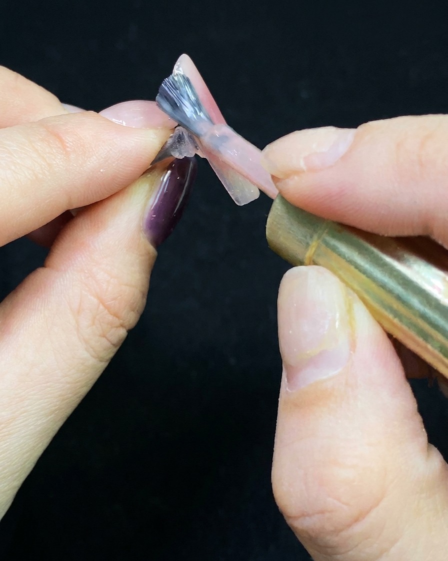 Un pincel aplicando una capa fina de esmalte gel nude sobre un tip transparente.