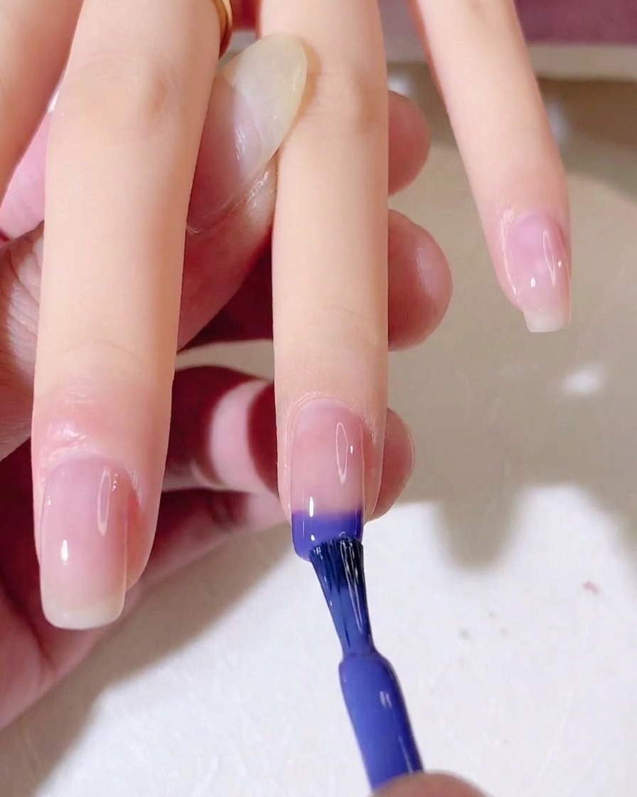 Un pincel de uñas aplicando esmalte de gel azul y morado en la punta de una uña natural.