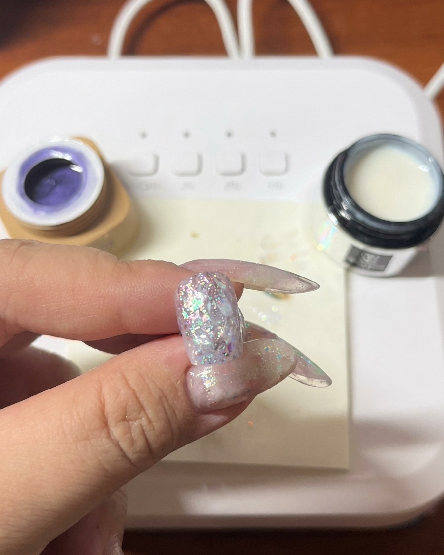 Un pincel aplicando gel morado sobre la uña ya decorada con glitter y conchas.