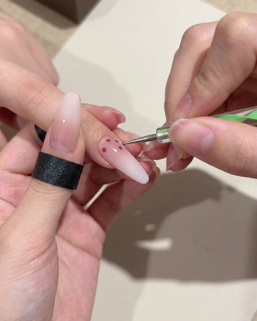 Primer plano de una herramienta de punteado aplicando pequeños lunares rojos precisos sobre una uña con base de degradado rosa suave.