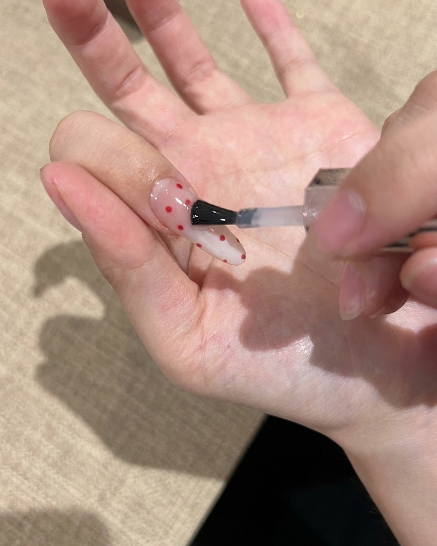 Primer plano de un pincel aplicando un top coat transparente y brillante sobre una uña decorada con lunares rojos.