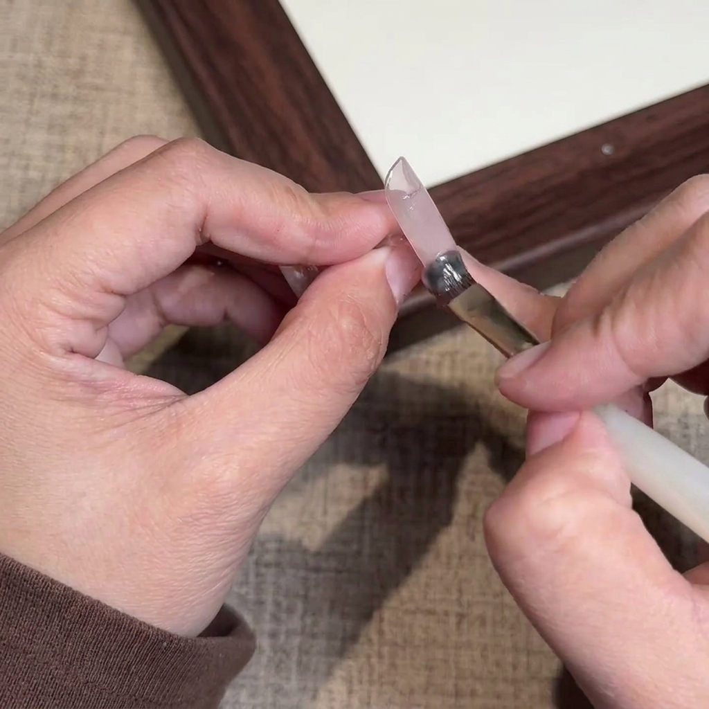 Primer plano de un pincel aplicando esmalte en gel rosa nude translúcido sobre una uña natural.