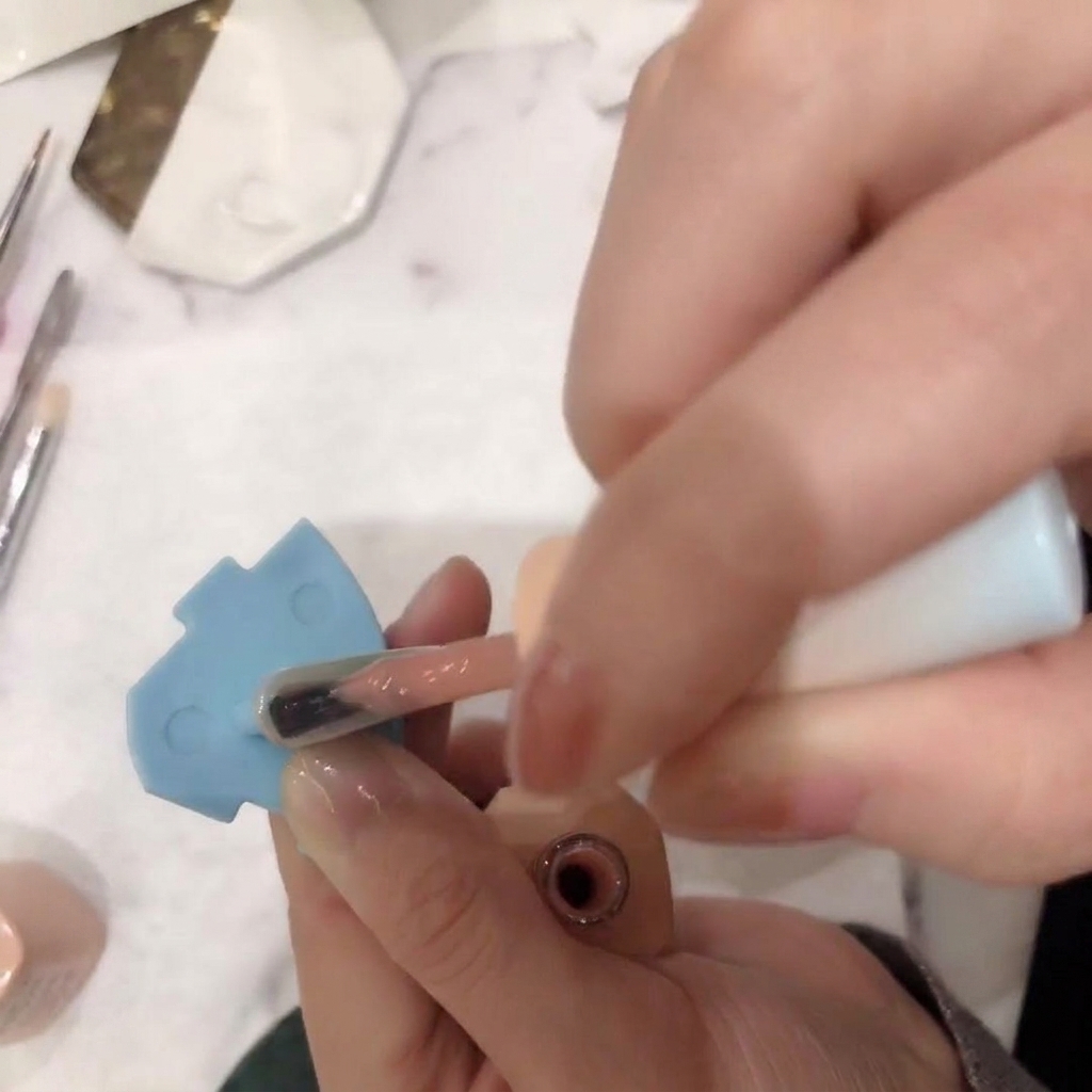 Primer plano de un pincel aplicando un esmalte en gel rosa nude traslúcido sobre la uña.