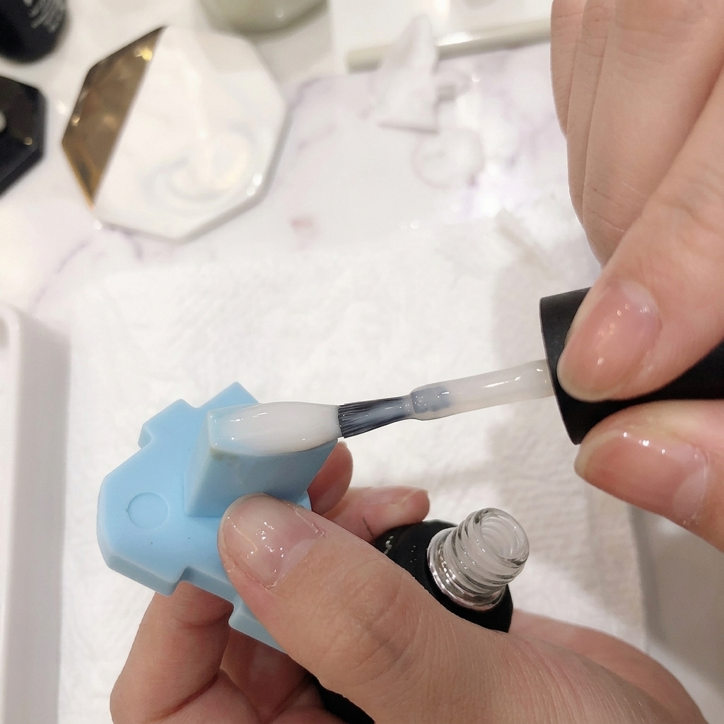 Primer plano de un pincel aplicando una capa traslúcida de esmalte blanco lechoso sobre una uña con ombré rosa y blanco.