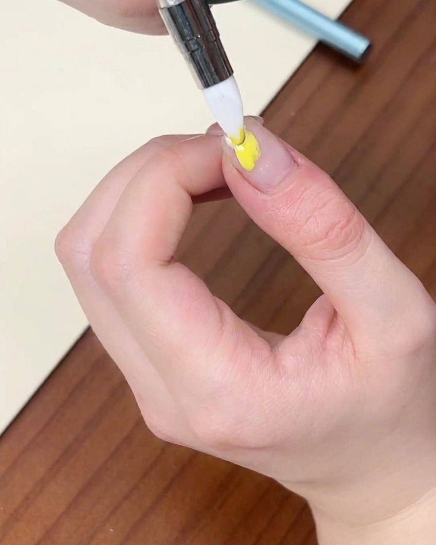 Un pincel fino aplicando una gota de esmalte en gel amarillo sobre una uña natural.
