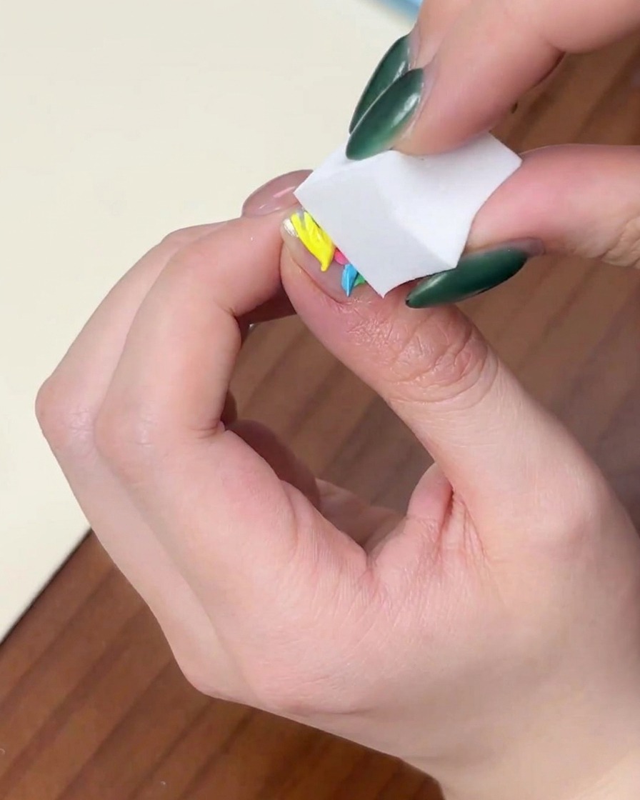 Primer plano de una mano presionando una esponja de maquillaje blanca verticalmente sobre una uña con gotas de esmalte en gel de colores.