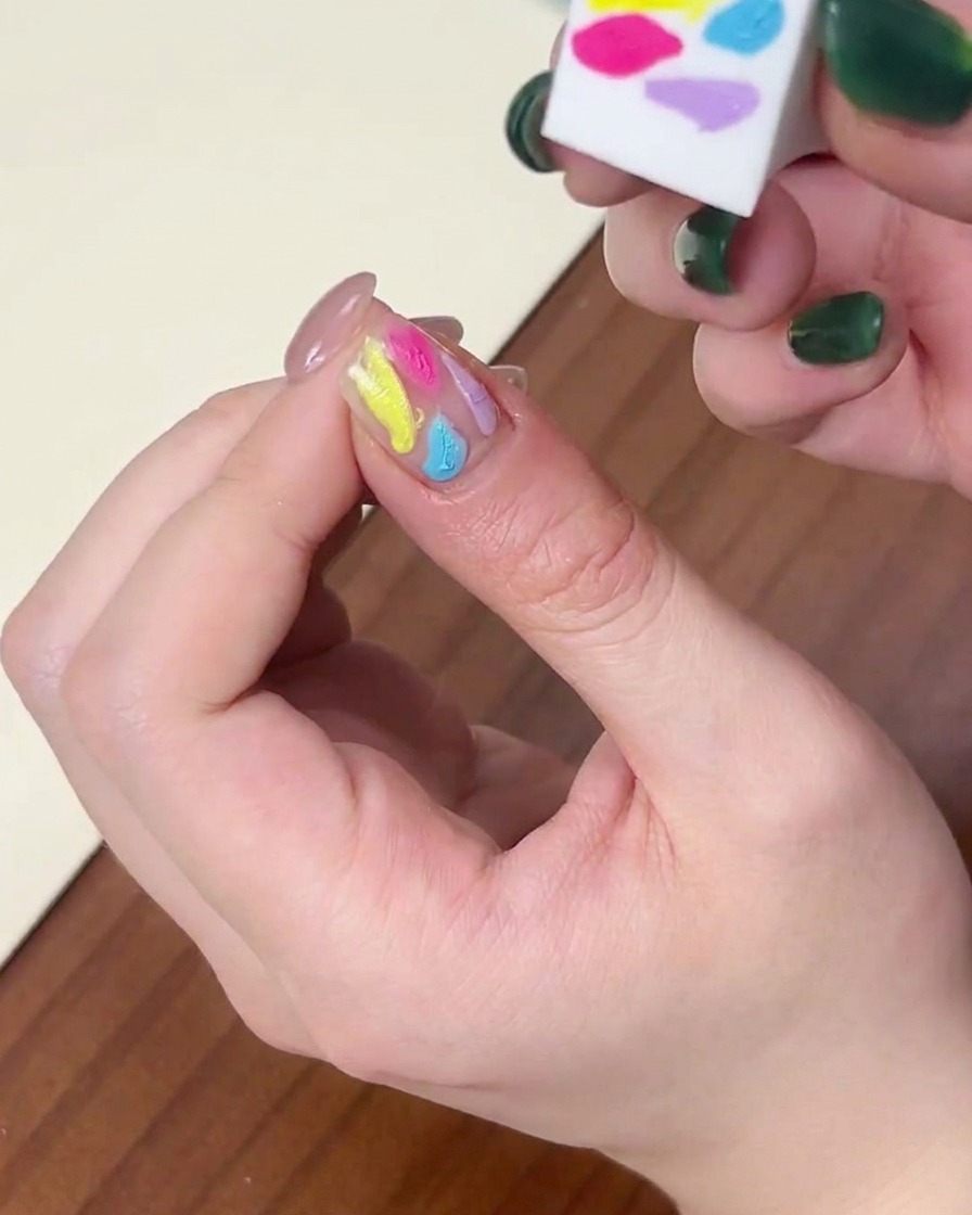 Primer plano del lateral de una esponja difuminando puntos multicolores de esmalte en gel sobre una uña.