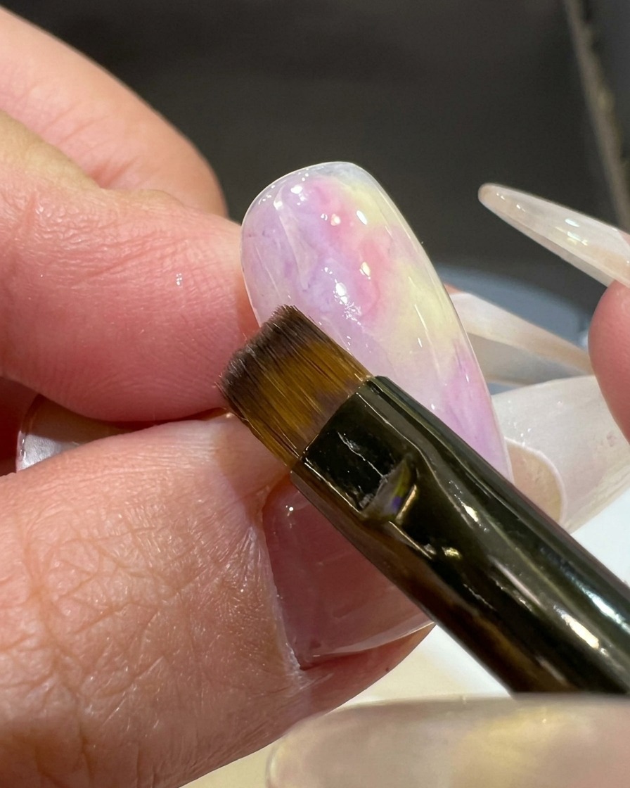 Un pincel plano aplicando un color de gel rosa violáceo traslúcido en la esquina de una uña con base blanca.