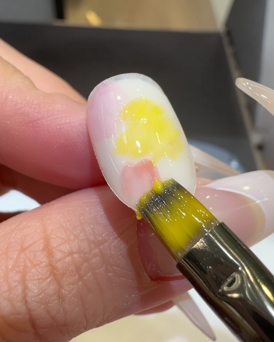 Un pincel plano mezclando esmalte de gel rosa y amarillo sobre la superficie de la uña.