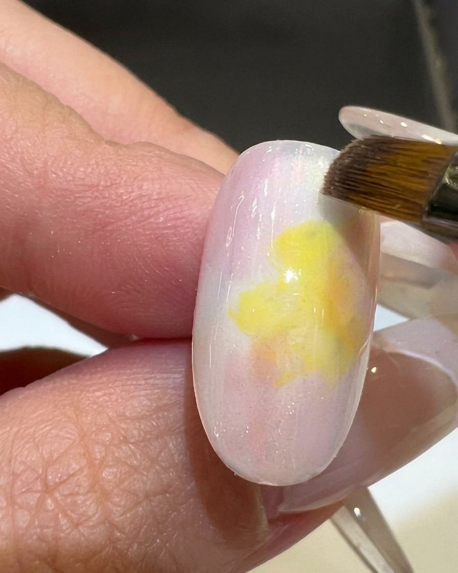 Un pincel plano suavizando el degradado entre el esmalte rosa y amarillo en una uña.