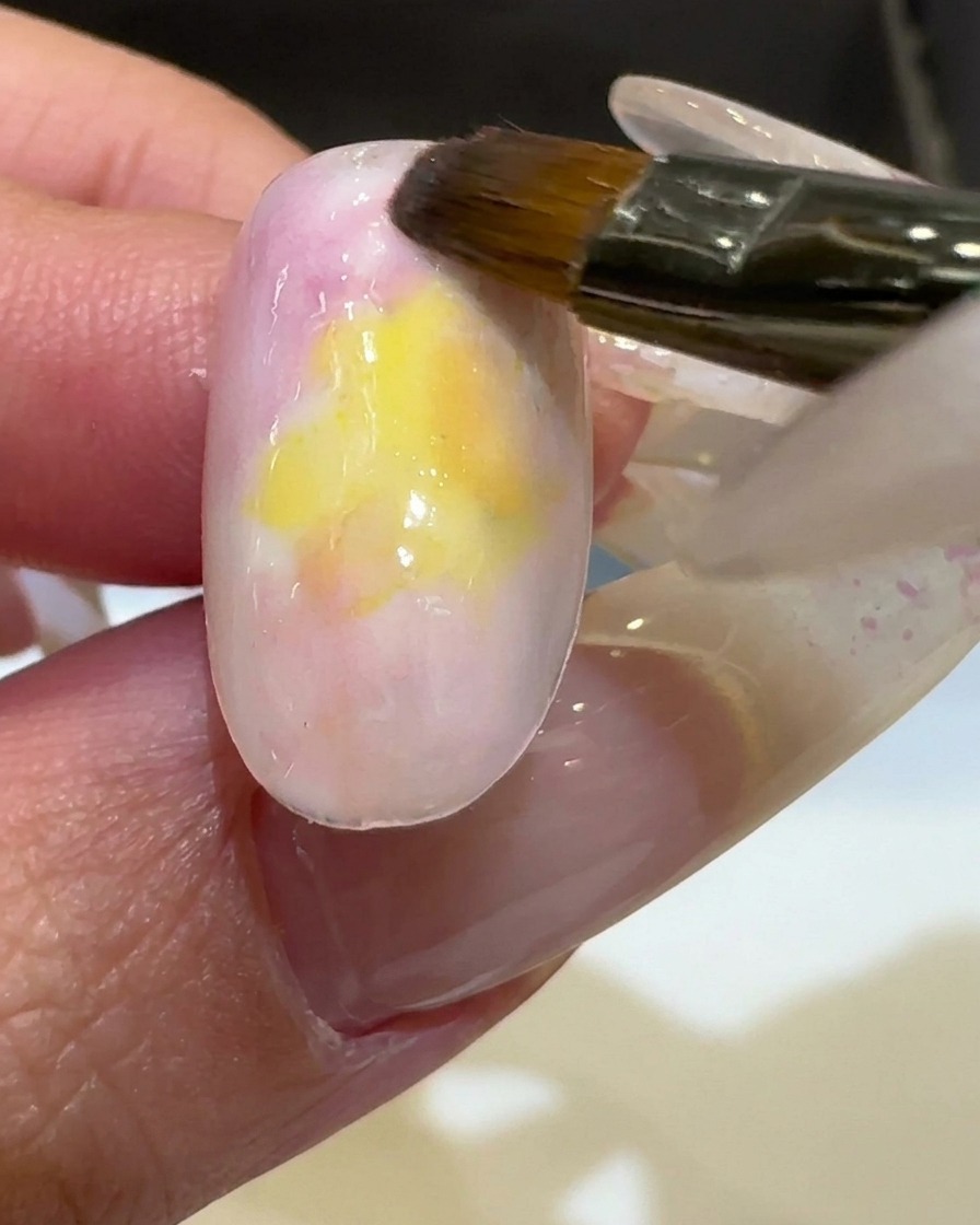 Un pincel aplicando un esmalte de gel amarillo vibrante en el centro de una uña, mezclándolo con los colores existentes.