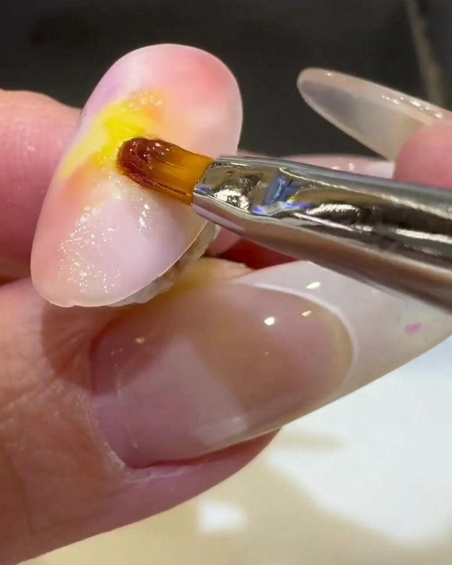 Un pincel delineador fino terminando una onda de gel 3D transparente en la mitad inferior de una uña color atardecer.