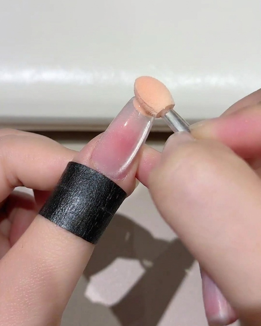 Un primer plano de una pequeña herramienta de esponja aplicando gel rosado en el borde libre de una uña transparente.