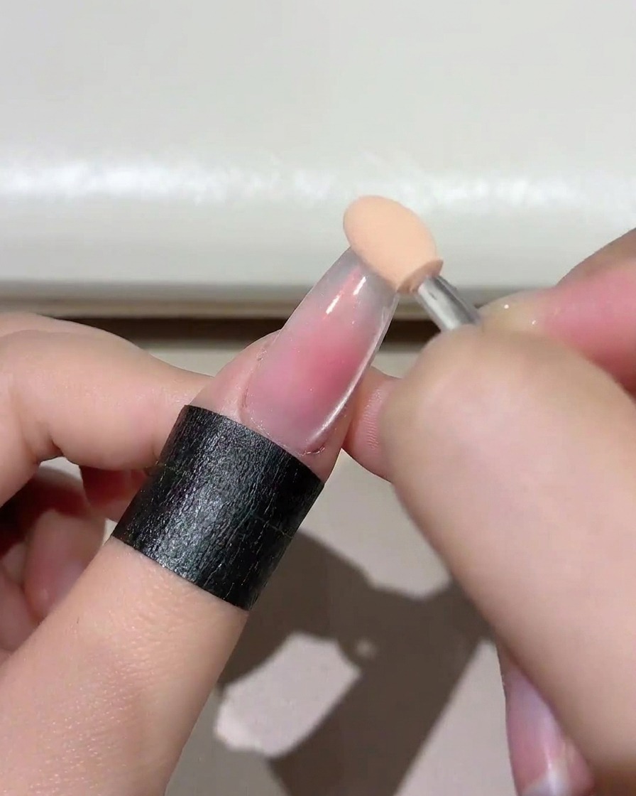 Un primer plano de un aplicador de esponja aplicando un gel rosa rubor suave sobre la punta de la uña transparente.