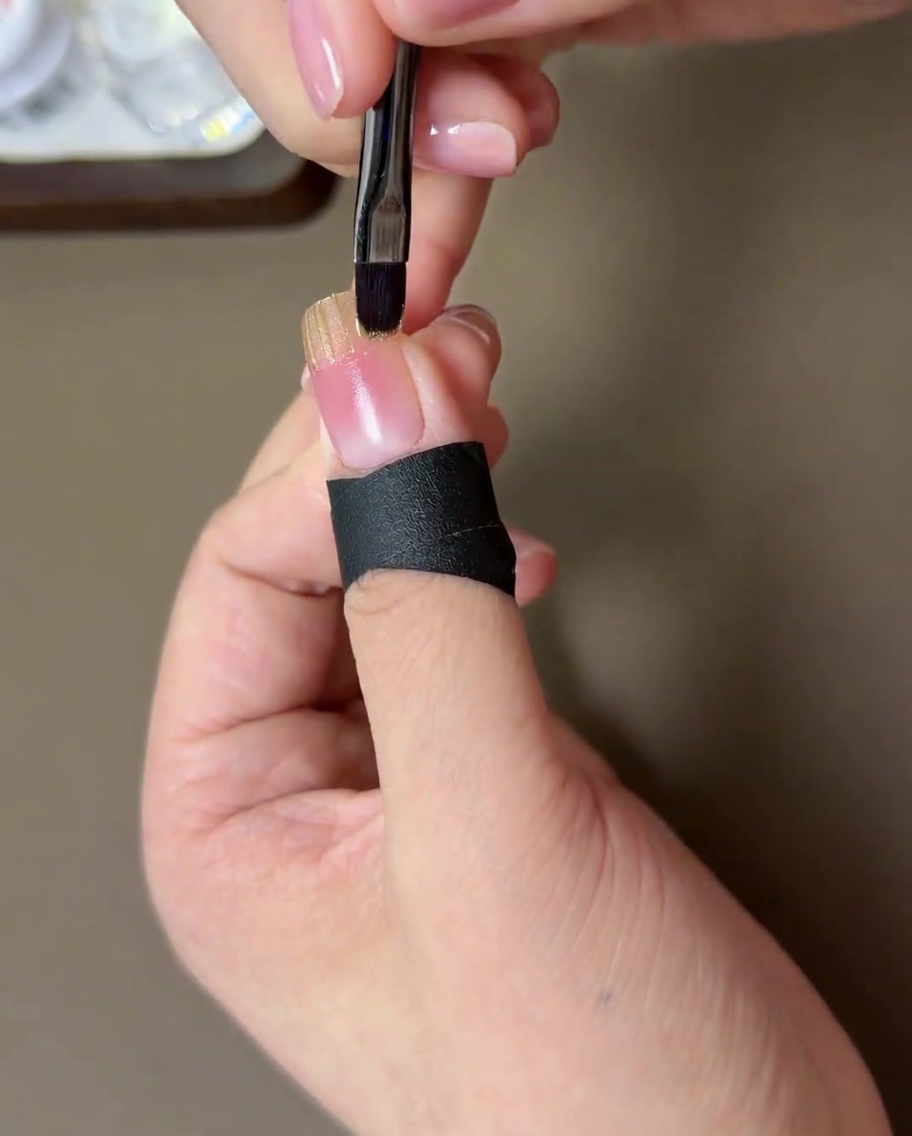 Un primer plano de un pincel pequeño difuminando el borde del gel dorado hacia la base rosa blush.
