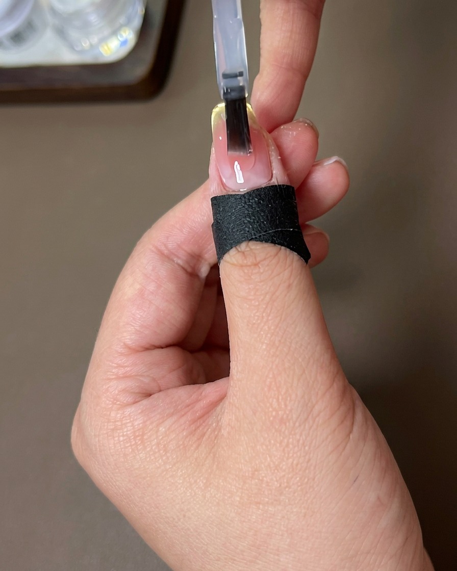 Un pincel aplicando una capa transparente y brillante de top coat sobre una uña con el degradado atardecer terminado y el efecto magnético francés.