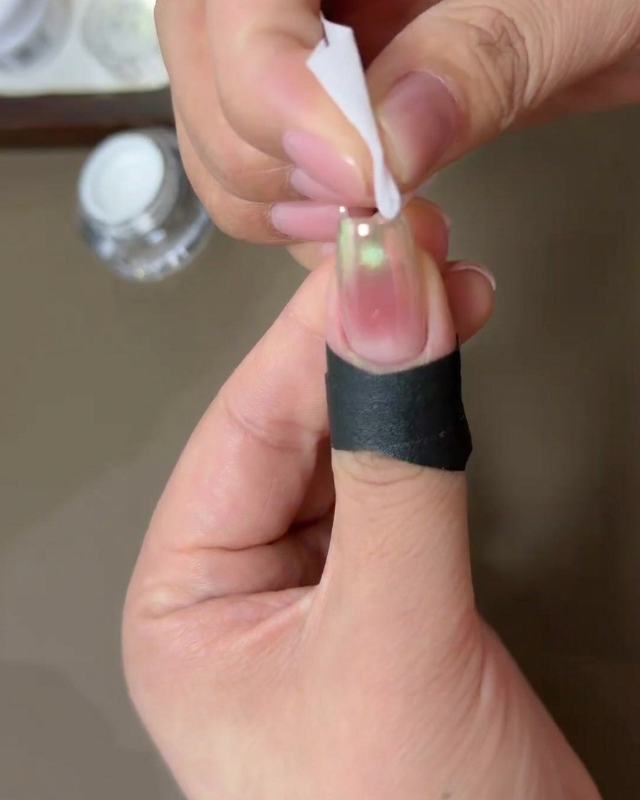 Una persona usando una pequeña toallita sin pelusa humedecida con alcohol para limpiar los bordes de una uña cubierta con polvo aurora chrome.