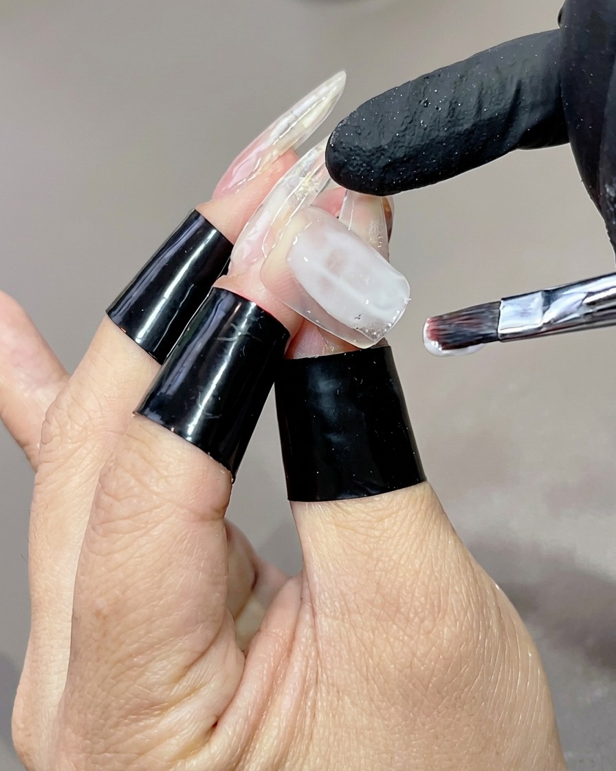 Una mano con tips transparentes largos, donde un pincel plano aplica una base de gel blanco lechoso en la uña del dedo anular.