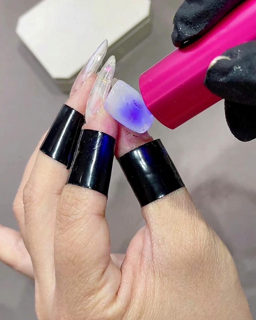 Primer plano de un dedo con una base de color blanco lechoso secándose bajo una pequeña linterna de mano LED o UV de color rosa.