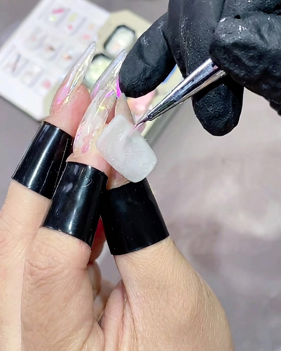 Un pincel fino aplicando un trazo horizontal de gel texturizado rosa de alto brillo sobre una uña pintada de blanco.