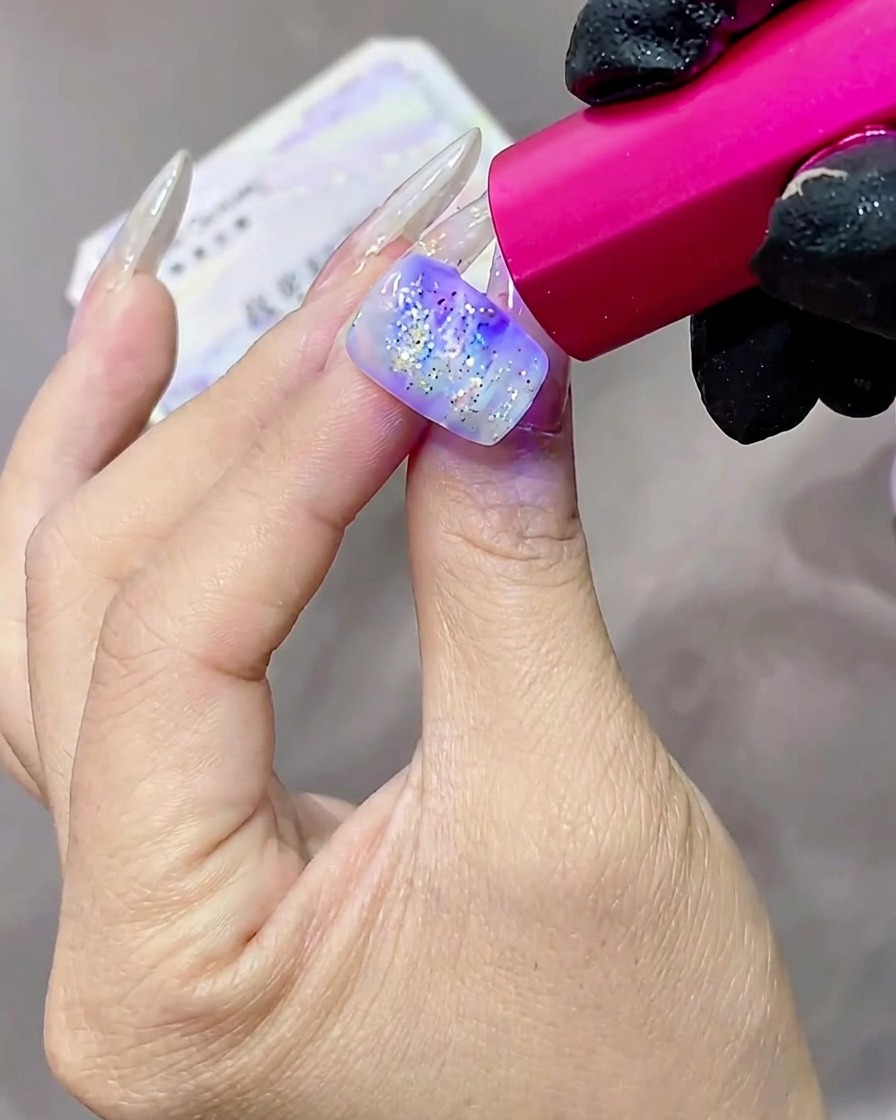 Una mano con uñas de gel texturizadas y con purpurina siendo curadas bajo una lámpara UV con tono rosado.
