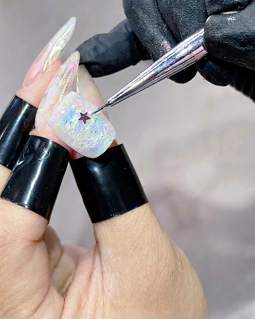 Una herramienta de metal colocando una pequeña lentejuela negra en forma de estrella en el centro de una uña cubierta con gel iridiscente y multicolor.