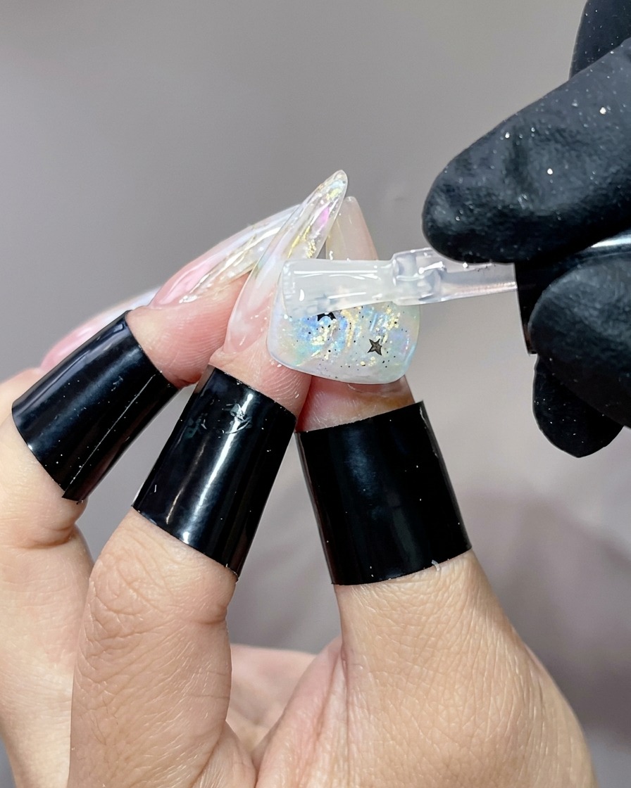 Un pincel aplicando una capa final de brillo transparente sobre un diseño de uña terminado con estrellas brillantes.